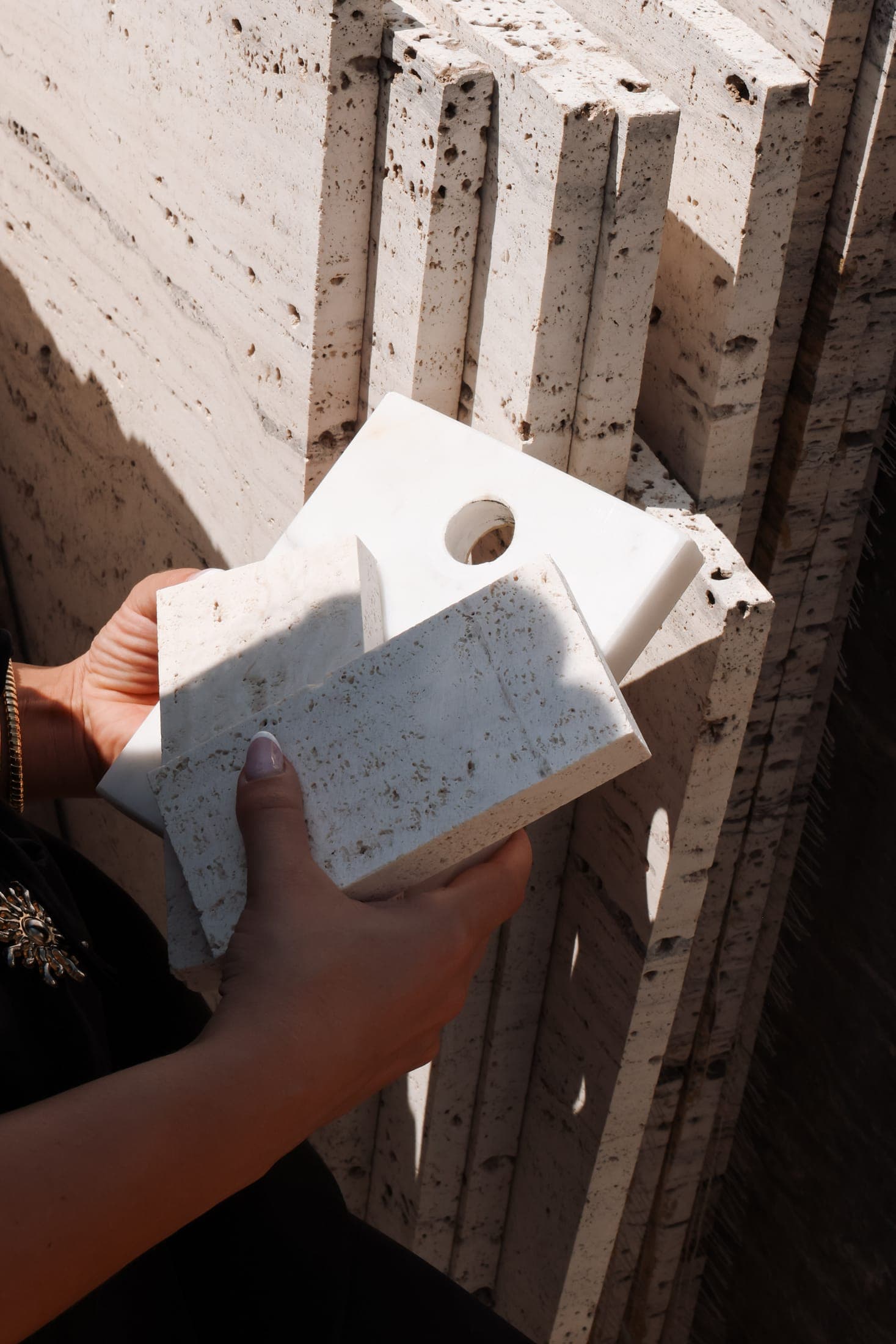 Travertine sample tiles held in hand in front of racked travertine slabs at the Zein Group yard